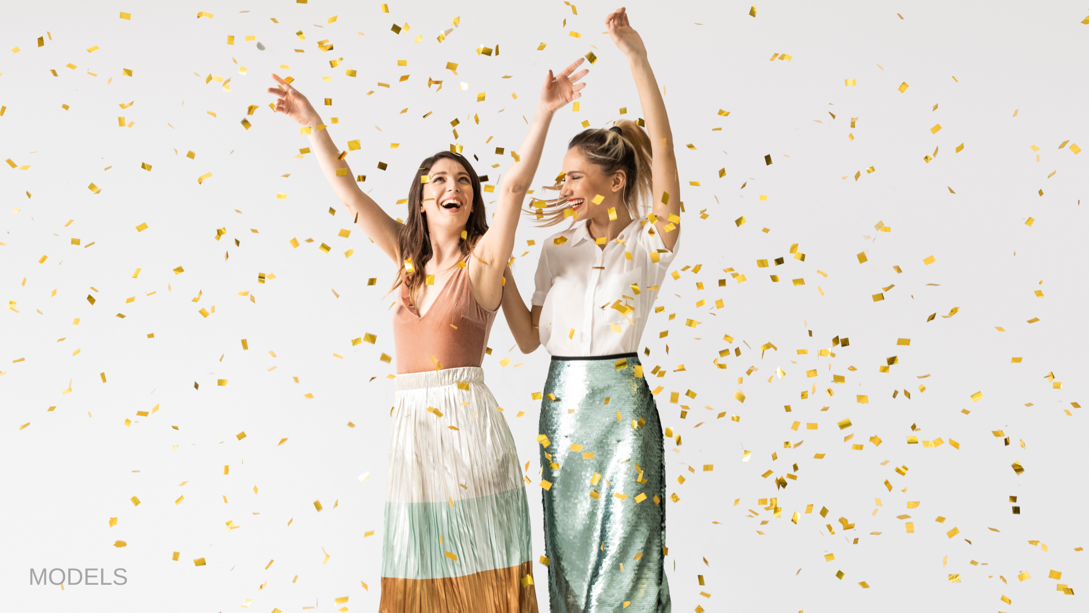 Two women standing in confetti with hands up and smiling. (MODELS)